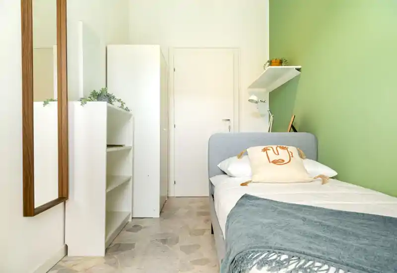 Camera singola compatta e luminosa con parete verde chiaro, letto singolo imbottito, plaid e cuscino decorativo, scaffalatura e specchio a tutta altezza. Stile pulito e moderno, ideale per un ospite singolo o affitti brevi.