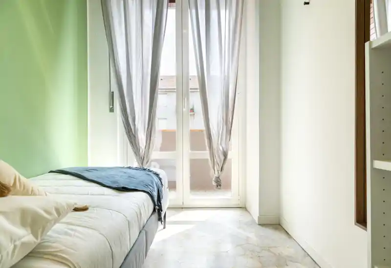 Camera da letto luminosa e semplice con letto singolo, parete verde chiaro e ampia porta finestra che lascia entrare molta luce naturale.