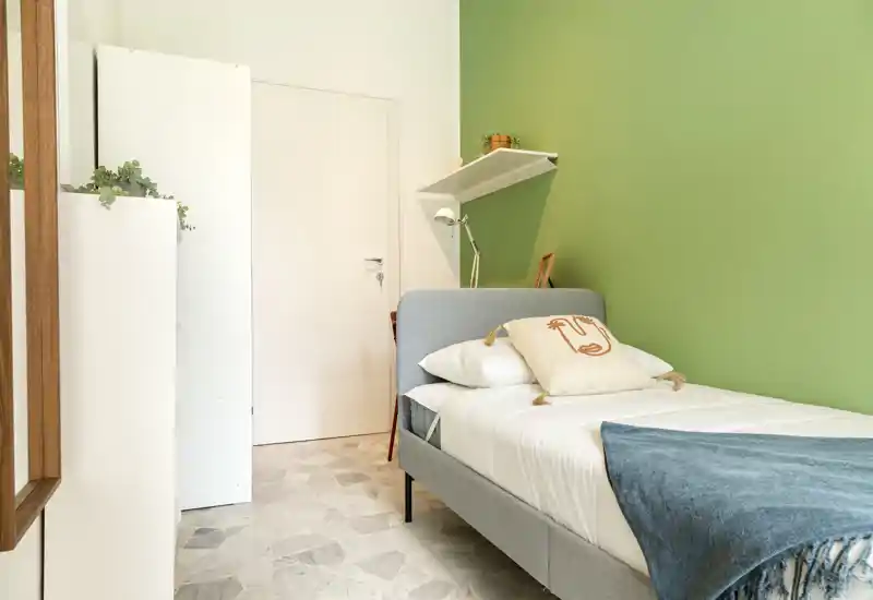 Camera da letto singola luminosa e ordinata con letto imbottito grigio contro una parete verde, biancheria bianca, cuscino decorativo e un plaid blu. La stanza è ben curata e illuminata, perfetta per un annuncio in affitto che punta su comfort e design essenziale.