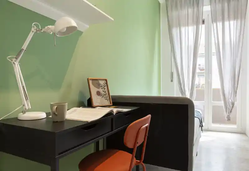 Angolo camera compatto con scrivania e letto parzialmente visibile accanto alla porta-balcone luminosa. La postazione ha una scrivania nera, lampada bianca regolabile e una sedia arancione che si abbinano alla parete verde e alle tende leggere.