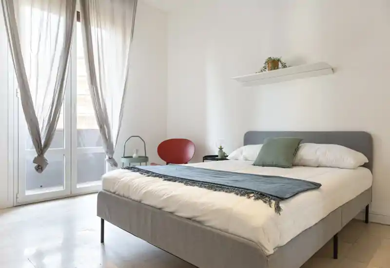 Dormitorio luminoso y minimalista con una cama doble cómoda, ropa de cama suave, una manta decorativa y luz natural de grandes ventanas — perfecto para una estancia relajante.