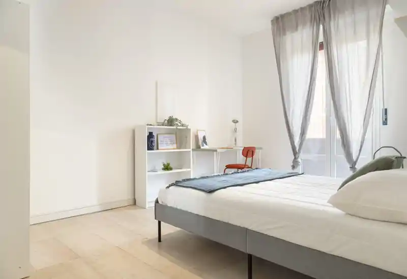 Dormitorio moderno y luminoso con cama doble bien hecha, textiles suaves y un pequeño escritorio junto a una gran ventana que aporta mucha luz natural.