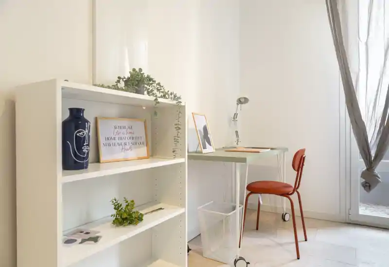Rincón del salón luminoso y minimalista con pequeña mesa de trabajo, estantería, plantas decorativas y marcos — un espacio de trabajo tranquilo y aireado.