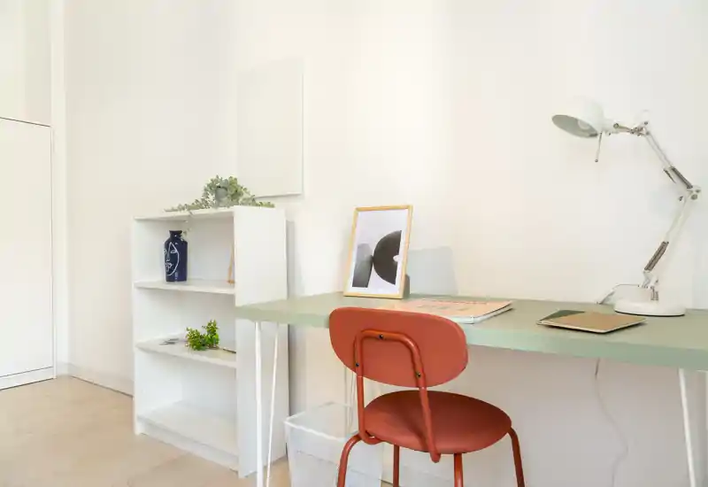 Rincón de trabajo luminoso y minimalista con mesa verde, silla roja, lámpara de escritorio regulable y una pequeña estantería blanca con objetos decorativos.