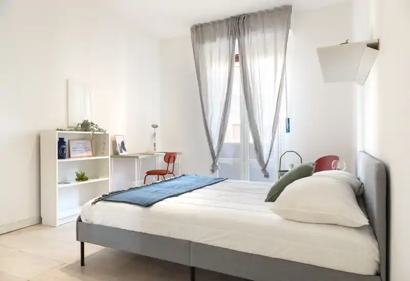 Chambre lumineuse et minimaliste avec un lit double bien fait, textiles doux et lumière naturelle provenant d'une grande fenêtre ; équipée d'un petit bureau et d'étagères pratiques.