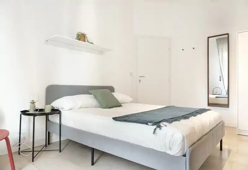 Chambre lumineuse et minimaliste avec un grand lit bien fait, de la literie douce, une petite table de chevet et un miroir mural — idéale pour une location moderne et paisible.