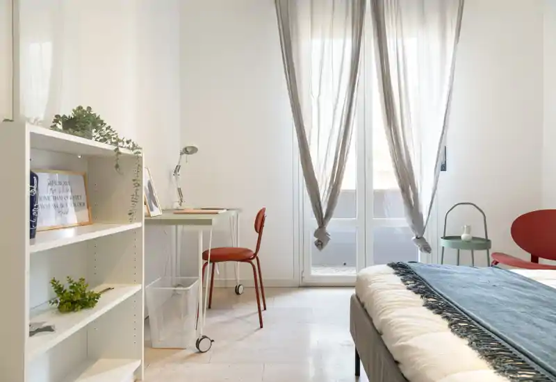 Chambre moderne et lumineuse avec lit bien fait, plaid bleu-vert, petit bureau et lumière naturelle provenant de grandes fenêtres de style français — parfaite pour un séjour confortable.