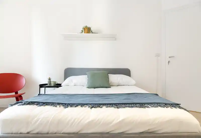 Chambre lumineuse et minimaliste avec un lit double central en linge blanc, plaid bleu et coussin vert ; table de chevet simple et décoration épurée.