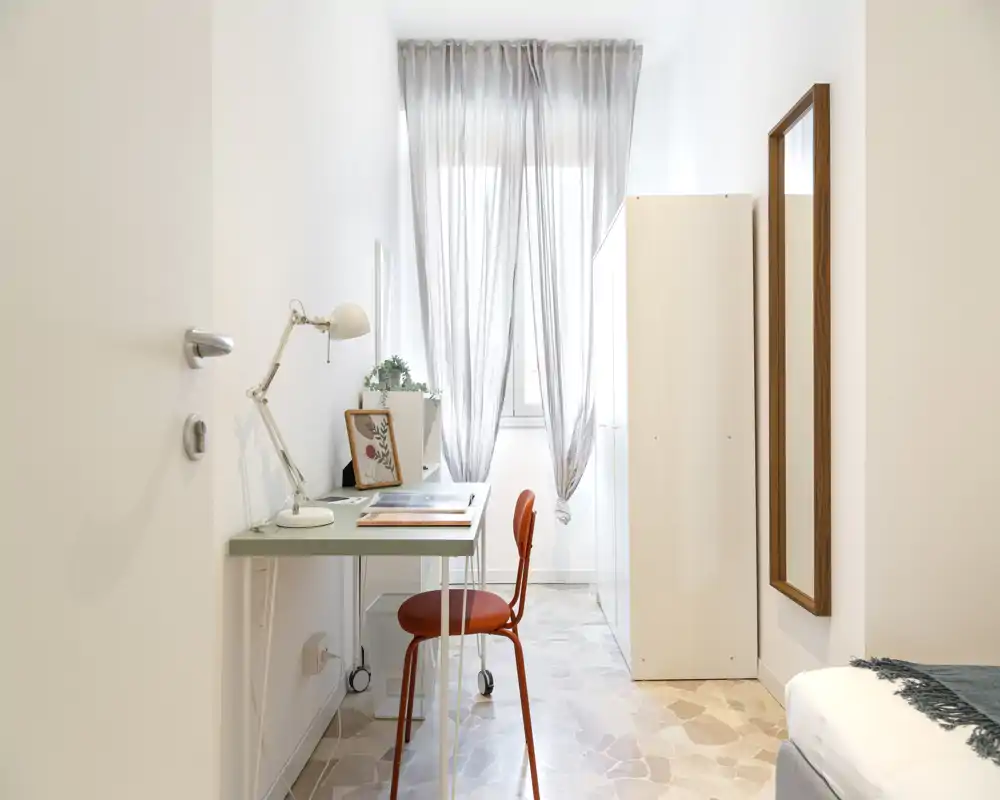 Compact, bright bedroom corner with a small desk and red chair beside a partially visible bed. The space features soft natural light, sheer curtains and minimal, modern decor ideal for a single occupant or short-term stay.