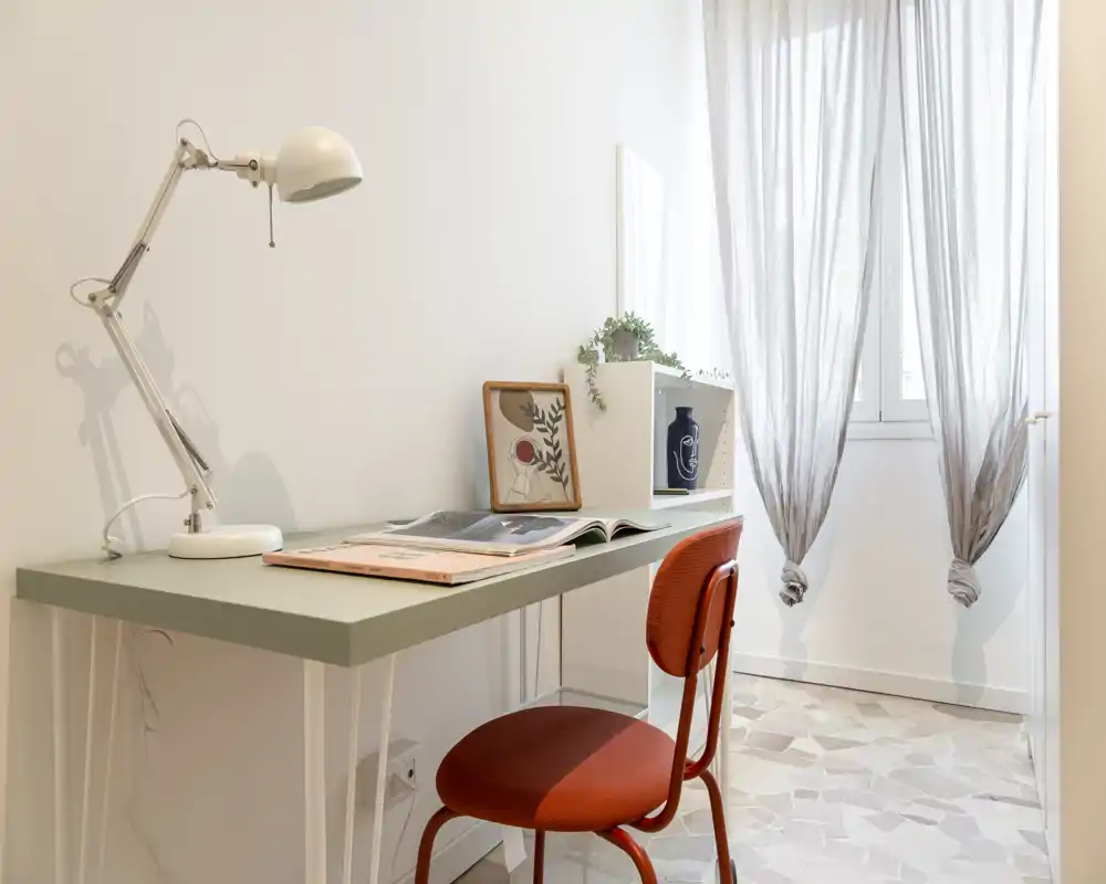 Bright, minimal desk area with a green tabletop, adjustable lamp and a warm red chair beside a window with sheer curtains — a cozy workspace corner in a small living area.