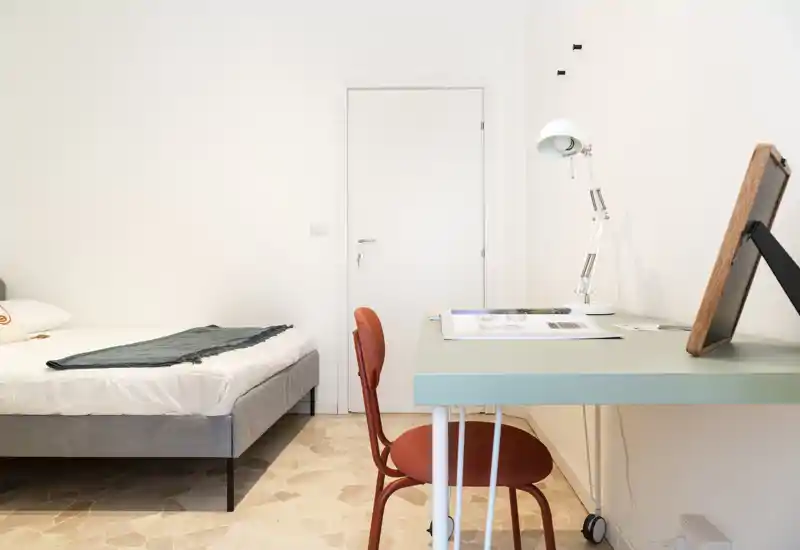 Minimal, bright bedroom with a low-profile double bed, neat white linens and a soft throw; a simple desk and chair create a functional workspace in the same room.