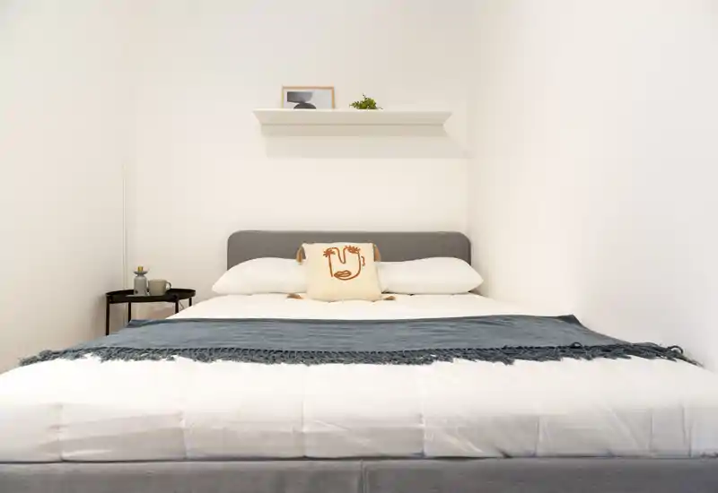 Bright, minimal bedroom with a centrally framed double bed, decorative pillow and a blue throw creating a calm, welcoming atmosphere.