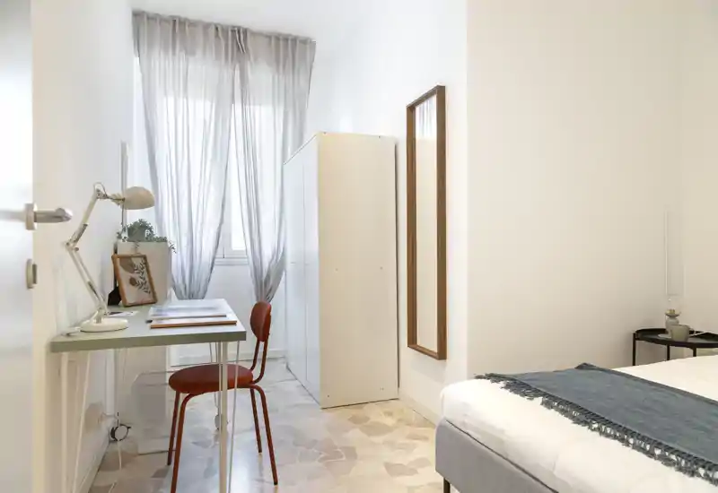 Bright, minimalist bedroom with a visible bed corner, soft textiles and a compact workspace by the window offering natural light.