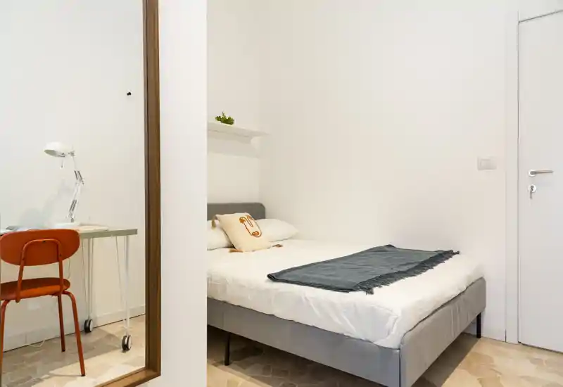 Bright, minimal bedroom with a comfortable double bed, textured throw and decorative cushion; a small desk and chair are visible in the reflection, making it ideal for a compact rental.