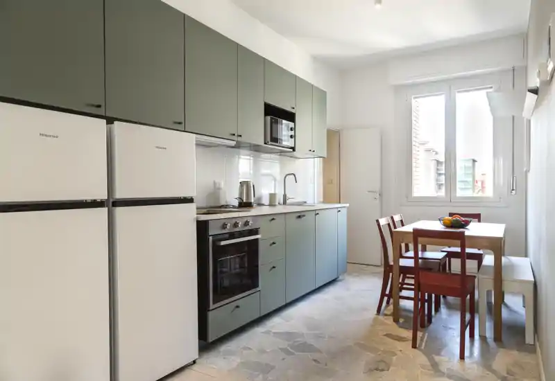 Cocina moderna y luminosa en línea con armarios verdes, horno empotrado, dos frigoríficos y una pequeña mesa de comedor junto a la ventana.