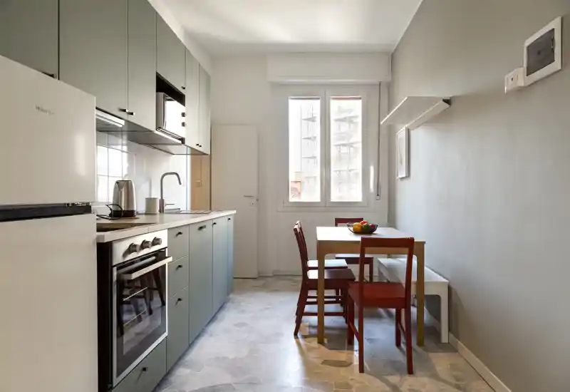 Cocina compacta y moderna con horno y nevera integrados, pequeña mesa de madera y cuatro sillas junto a una ventana luminosa — diseño limpio y funcional, ideal para un piso en la ciudad.