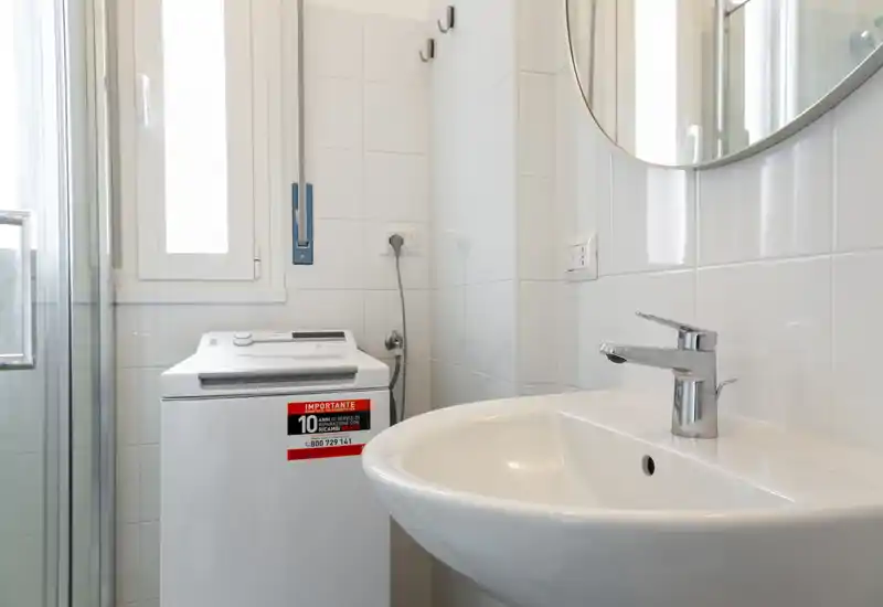 Rincón de baño compacto y luminoso con lavabo blanco y grifo cromado, espejo circular en la pared y una lavadora de carga superior junto a una pequeña ventana.