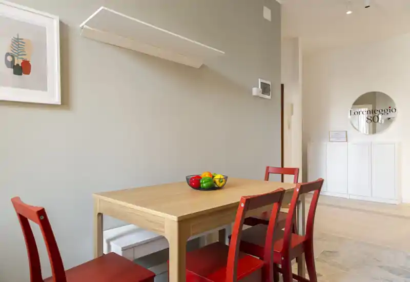 Rincón de comedor moderno y luminoso con mesa de madera clara, cuatro sillas rojas y un cuenco de frutas decorativo — minimalista, bien iluminado y acogedor para comidas informales.