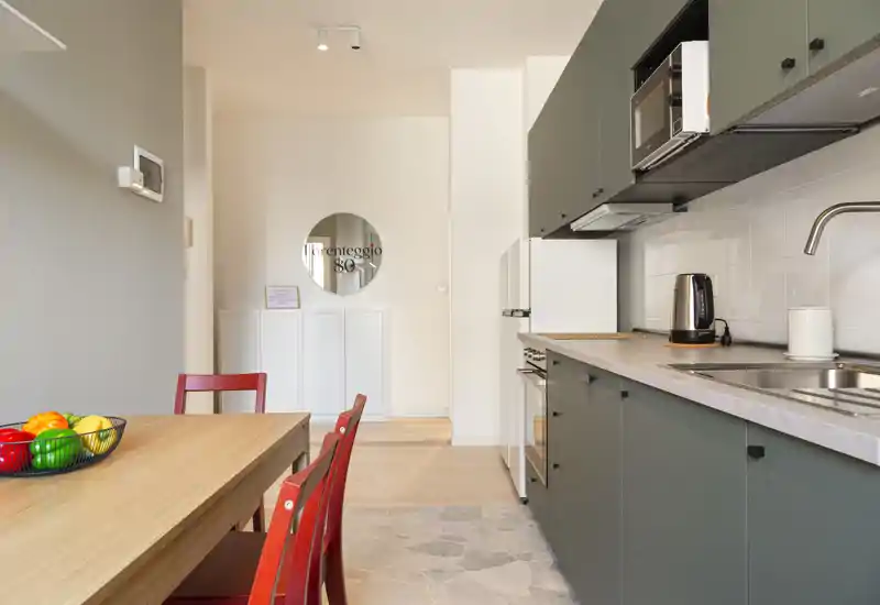 Cocineta moderna con armarios verdes, fregadero integrado, microondas y mesa de madera con sillas rojas: espacio luminoso y acogedor, ideal para alquileres de corta estancia.