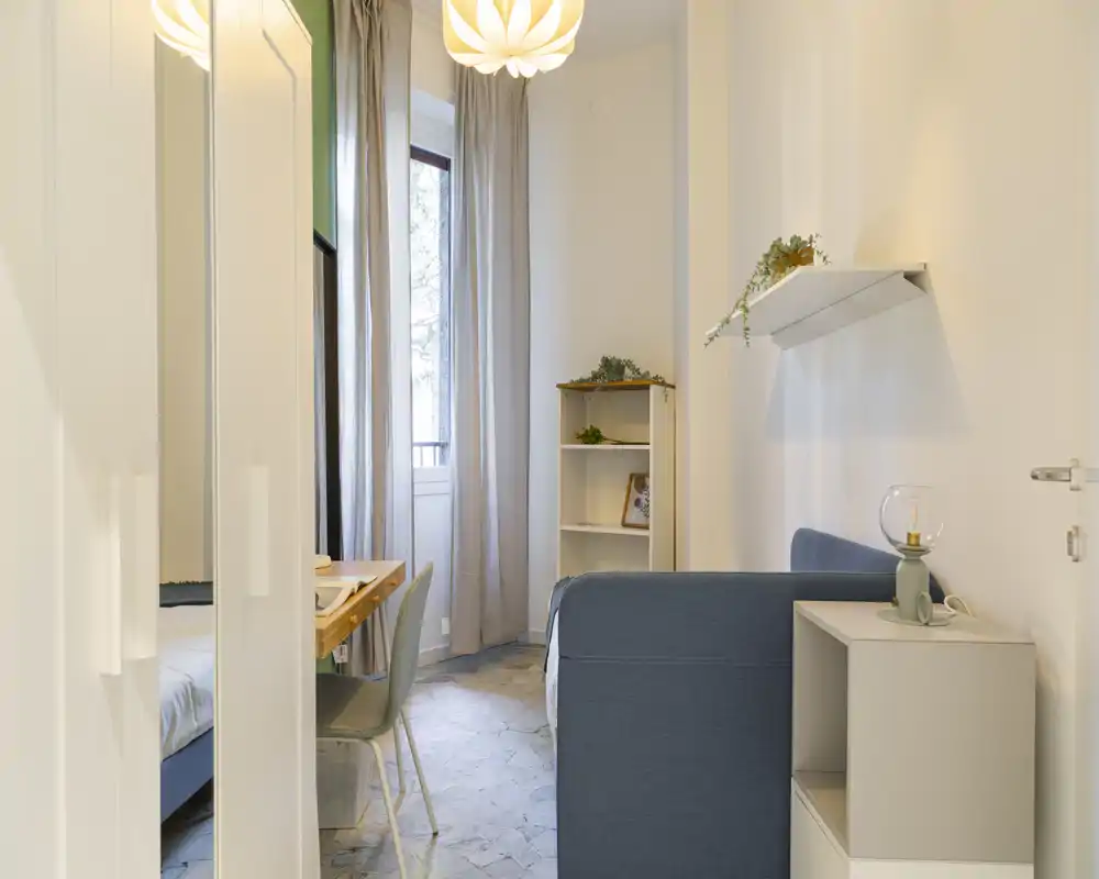 Compact, well-styled bedroom with a visible double bed, a small desk by the window, shelving and soft natural light creating a calm atmosphere.