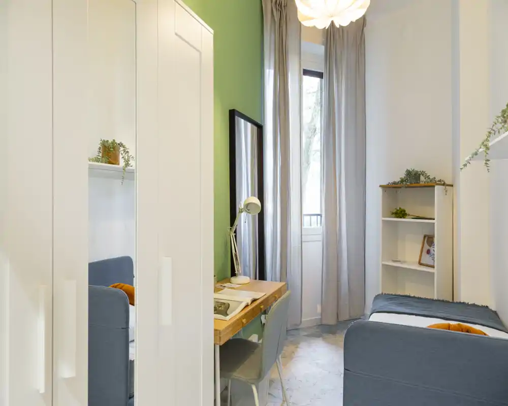 Bright, well-styled single bedroom with two compact beds, a wooden desk and wardrobe; neutral tones with green accent wall and natural light from a tall window.