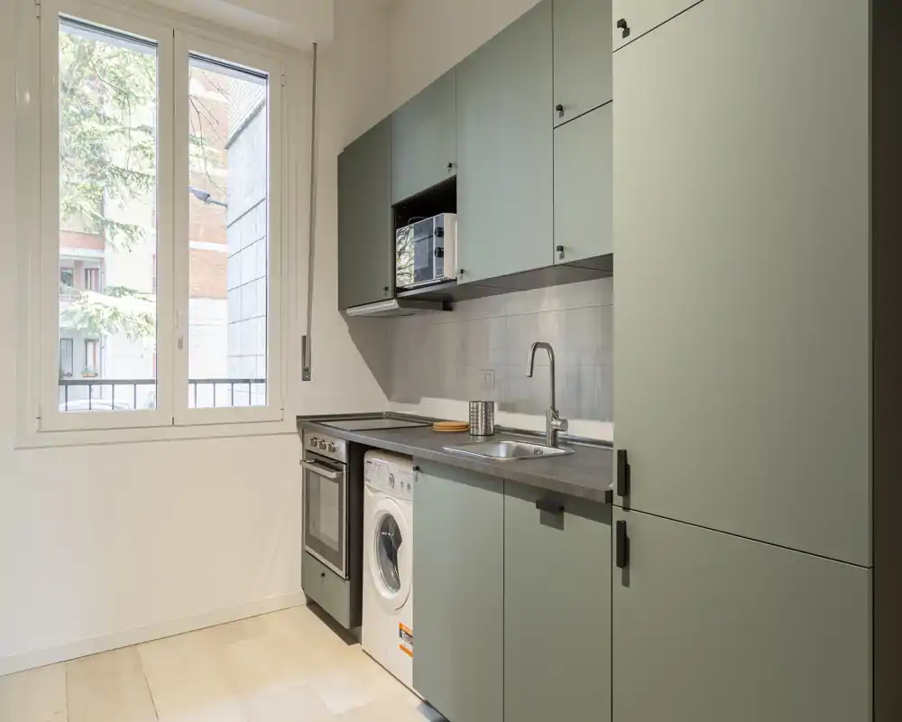 Bright compact kitchen with sage-green cabinetry, integrated appliances and a large window providing natural light. Functional layout includes sink, oven and a built-in washing machine.