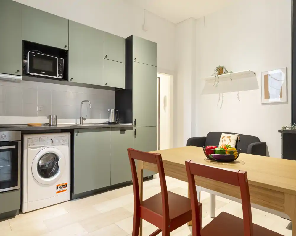 Bright modern kitchen corner with green cabinets, integrated appliances and a wooden dining table; cozy details include a fruit bowl, a small sofa and wall shelf plants.
