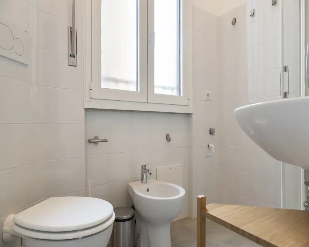 Clean, well-lit bathroom with a toilet, bidet, wall-mounted sink and frosted window; neutral tiles and simple wooden shelf create a fresh, minimalist feel.