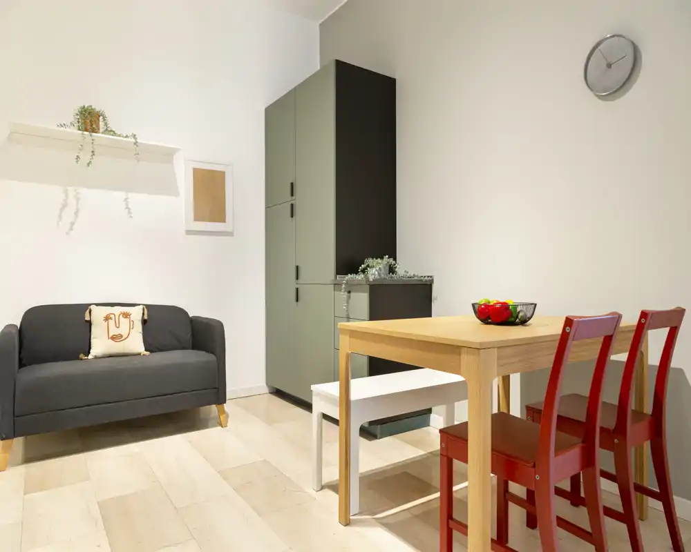 Bright, neatly staged living/dining area with a compact sofa, wooden dining table and red chairs; modern storage cabinet and small decorative plants create a tidy, welcoming space.