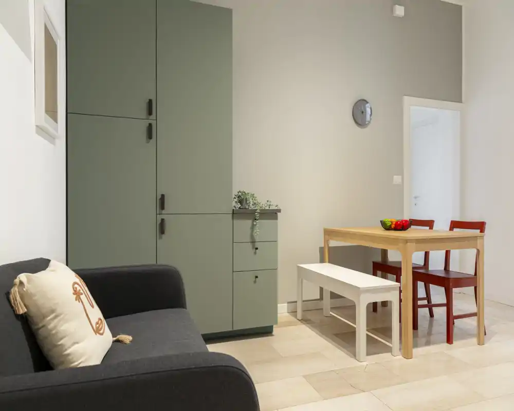 A compact, stylish living/dining corner with a dark sofa, green storage unit and a small wooden dining table with bench and red chairs; softly lit and neatly staged for a cozy, modern feel.