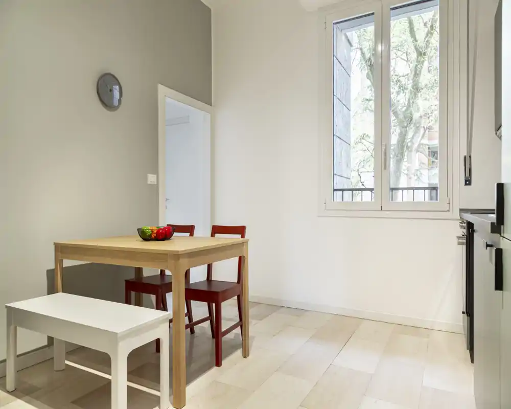 Minimal, bright dining area with a wooden table, two red chairs and a bench beneath a large window; simple, modern styling and natural light make it ideal for small meals.