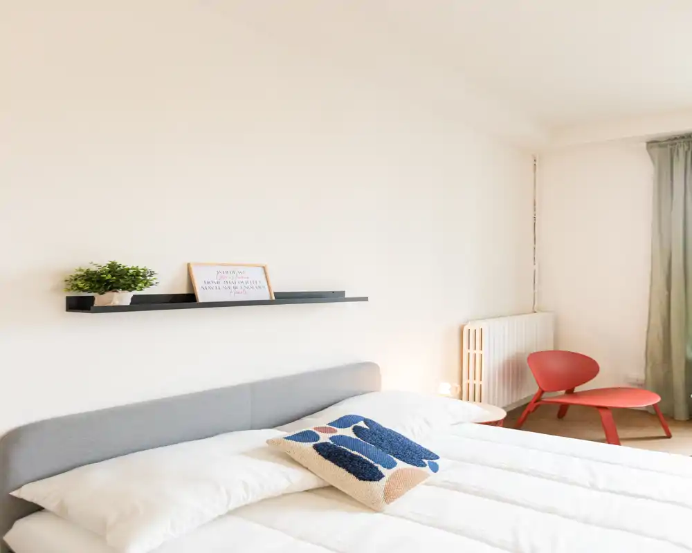 Camera da letto luminosa e minimal con letto matrimoniale ben rifatto, cuscino decorativo, mensola a parete con pianta e stampa incorniciata e una sedia rossa nell'angolo.