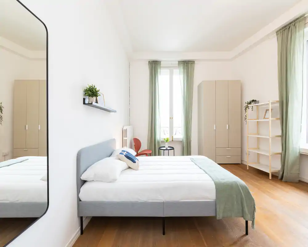 Camera da letto luminosa e moderna con un grande letto imbottito al centro, tessili morbidi e luce naturale da due alte finestre; toni neutri e scaffalatura minimal creano uno spazio calmo e accogliente.