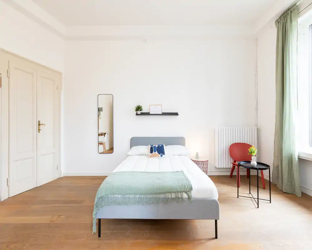 Camera da letto luminosa e minimal con un letto matrimoniale ben rifatto al centro, luce naturale da una grande finestra e arredi semplici come specchio, mensola e tavolino.