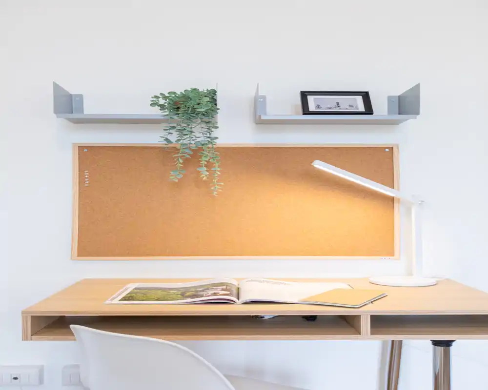 Angolo scrivania luminoso e minimal con bacheca in sughero, lampada, pianta sospesa e libro aperto — ideale come ufficio domestico o angolo studio.