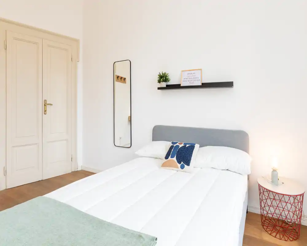 Camera da letto luminosa e minimalista con letto matrimoniale ben rifatto, testiera semplice, lampada da comodino e cuscino decorativo; pareti bianche e pavimento in legno creano un'atmosfera calma e moderna.