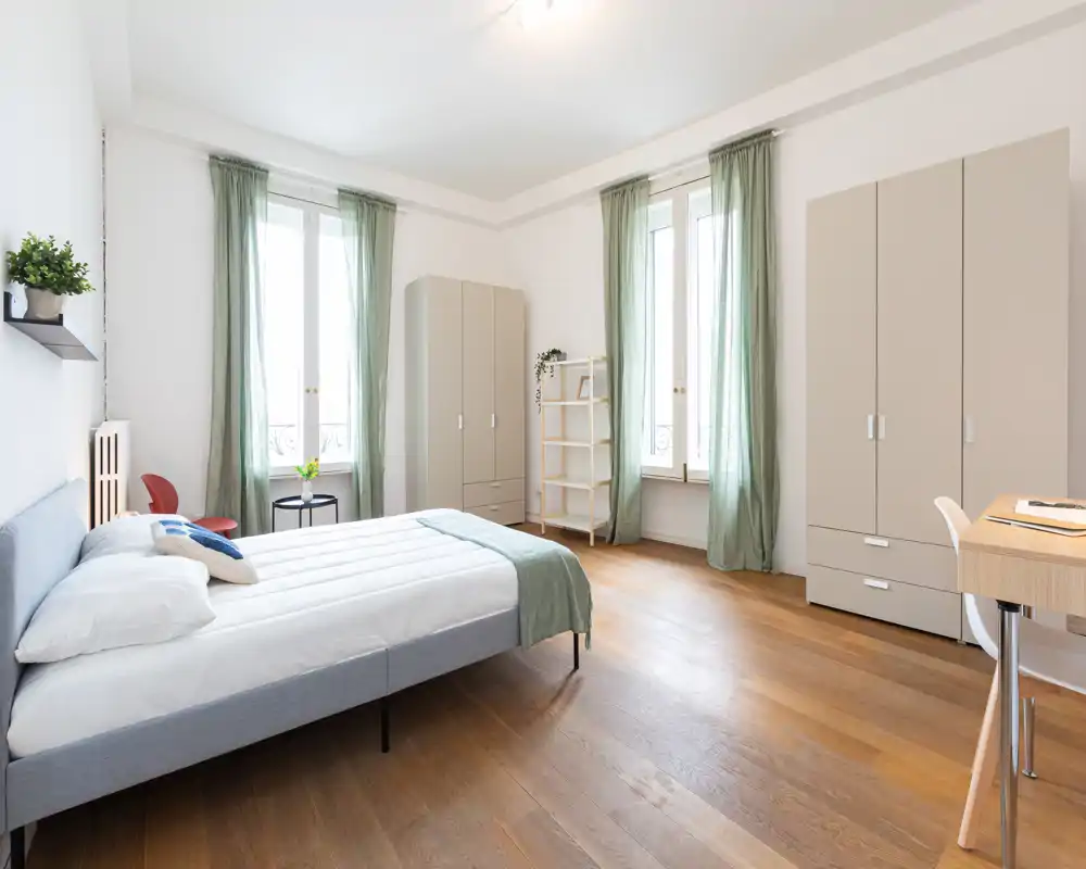 Camera da letto luminosa e ariosa con un ampio letto matrimoniale, pavimento in legno e alte finestre con tende verde chiaro. Armadio moderno e scrivania minimal completano lo spazio rilassante.