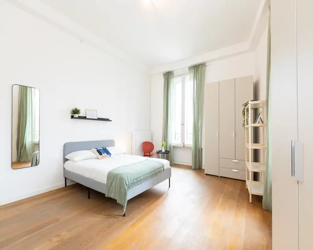 Camera da letto luminosa e minimal con letto matrimoniale, tocchi verdi e luce naturale da una finestra alta; pavimenti in legno chiaro e ampio armadio creano un ambiente arioso e rilassante.