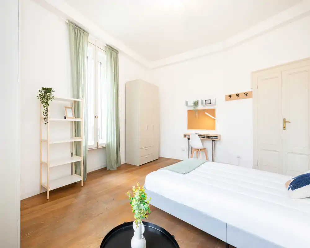 Camera da letto luminosa e minimal con letto ben rifatto in primo piano, armadio e scrivania vicino alla parete. Ampia finestra con tende verde chiaro che dona molta luce naturale e pavimento in legno che crea calore.