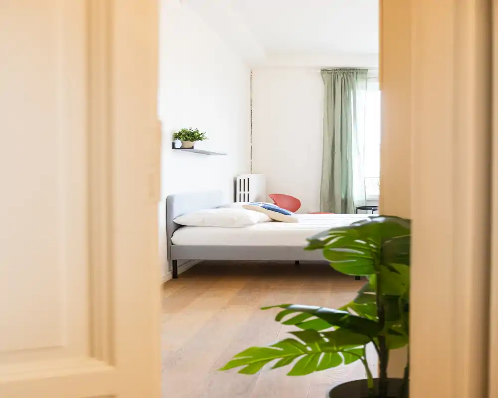 Camera che inquadra una camera da letto luminosa attraverso una porta: letto basso grigio con cuscini, pavimento in legno chiaro, tende verdi; cuscini decorativi e una pianta aggiungono calore.