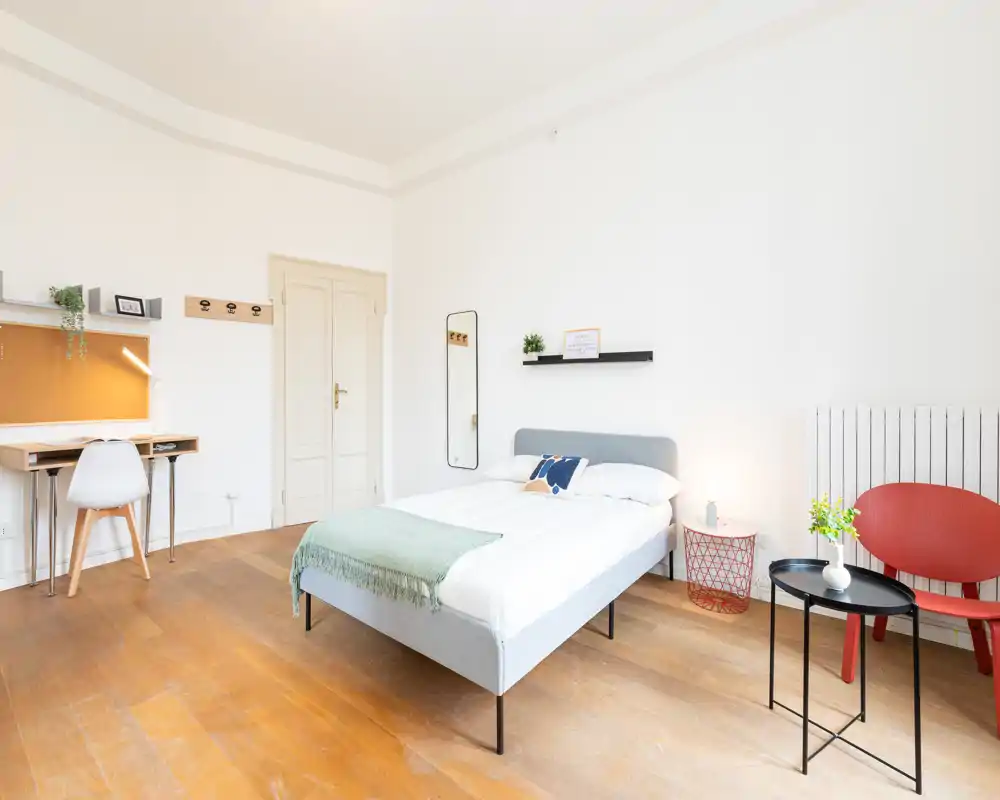Camera da letto luminosa e minimal con letto matrimoniale ben fatto, piccolo comodino e scrivania; pavimento in legno e toni neutri creano un'atmosfera rilassante.