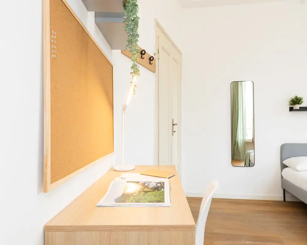 Angolo camera da letto luminoso e minimalista con scrivania in legno, bacheca in sughero e letto grigio parzialmente visibile — stile nordico pulito adatto a un annuncio.