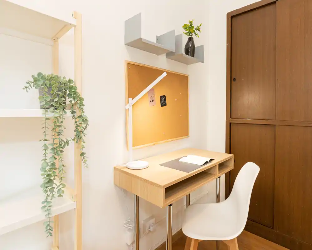 Angolo studio compatto e ben illuminato con scrivania in legno, sedia bianca, bacheca in sughero e mensole minimaliste — ideale per lavoro o lettura concentrata.