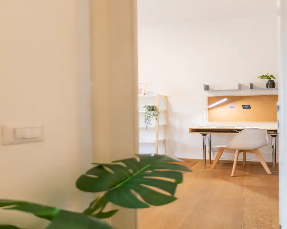 Angolo studio luminoso e minimal con scrivania in legno, bacheca in sughero e una sedia bianca moderna; piante e scaffali donano un tocco accogliente.