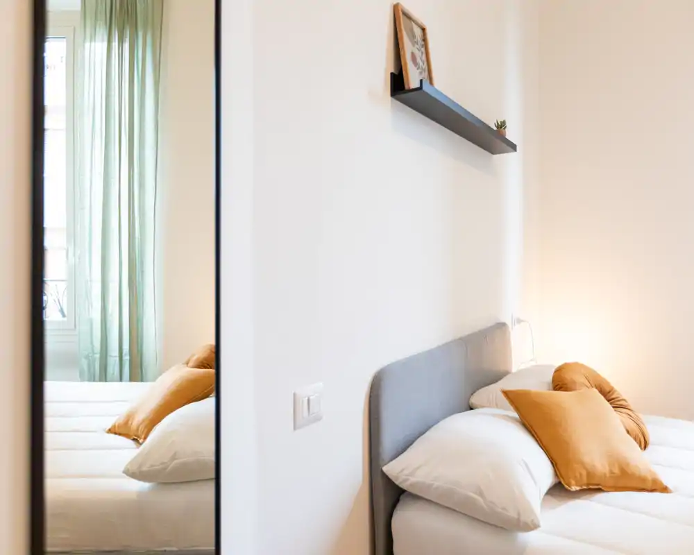 Camera da letto luminosa e ben curata con testiera grigio chiaro, cuscini ocra caldi e biancheria a strati; la luce naturale filtrata dalla tenda crea un’atmosfera rilassante.