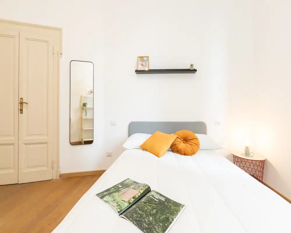 Camera da letto luminosa e minimal con un letto ben rifatto, cuscini colorati, lampada da comodino e una mensola semplice: uno spazio moderno e tranquillo perfetto per riposare.