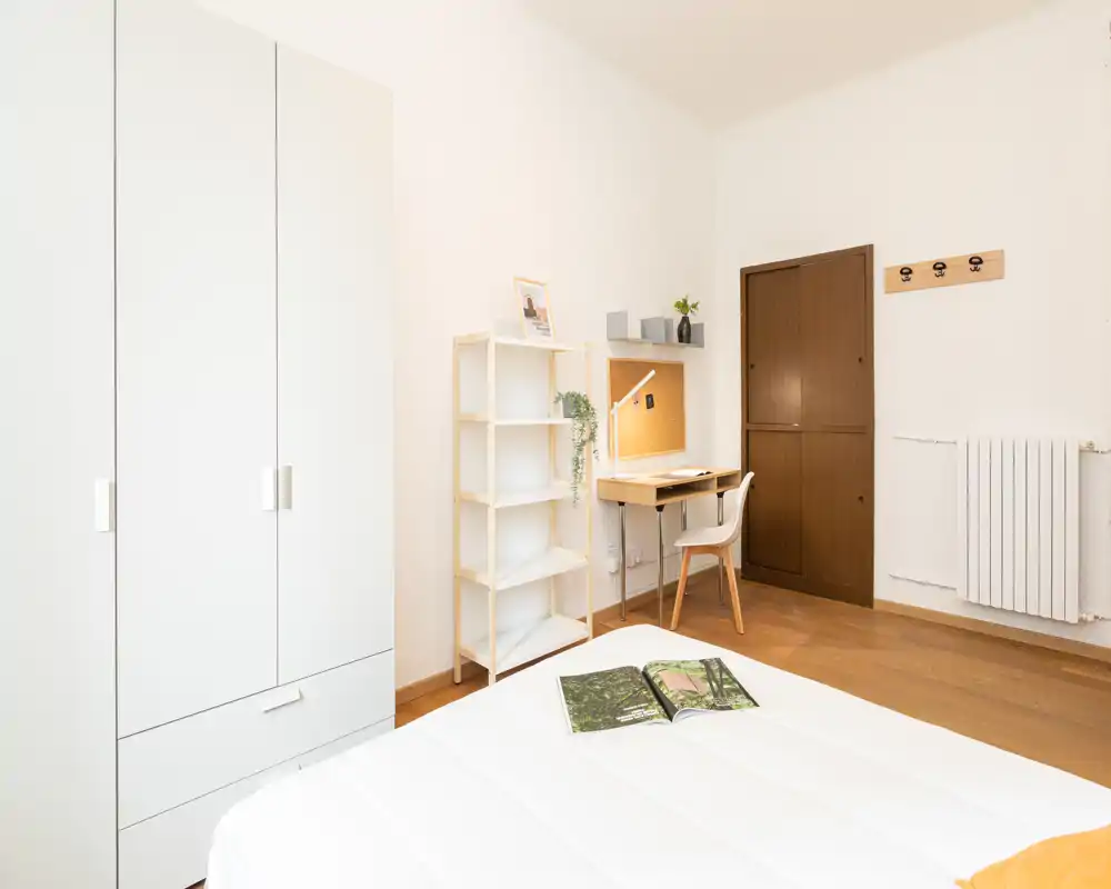 Camera da letto luminosa e minimale con letto ordinato in primo piano, armadio bianco, scaffalatura e una piccola scrivania in legno con sedia — pulita, moderna e ben illuminata.