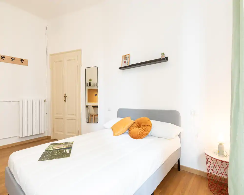 Camera da letto luminosa e minimal con letto imbottito grigio, cuscini colorati caldi e pavimento in legno; mensola semplice e lampada da comodino creano un aspetto accogliente e moderno.