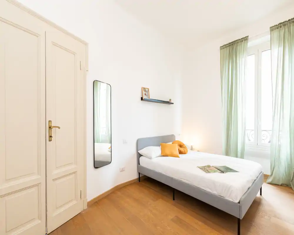 Camera da letto luminosa e minimal con letto matrimoniale, testiera grigia, tende verdi leggere e pavimento in legno caldo — perfetta per un soggiorno accogliente in città.