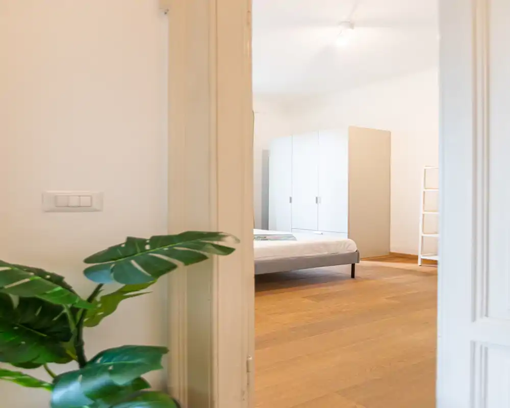 Camera da letto luminosa e minimal vista attraverso una porta aperta, con letto basso, armadio bianco e pavimenti in legno caldo che creano un'atmosfera tranquilla e contemporanea.